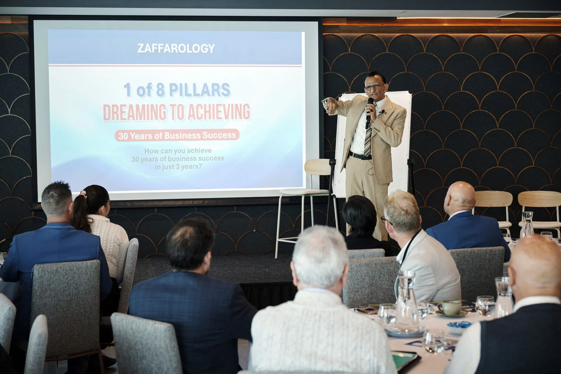 Zaffar Khan pointing to the audience beside a slide reading Dreaming to Achieving