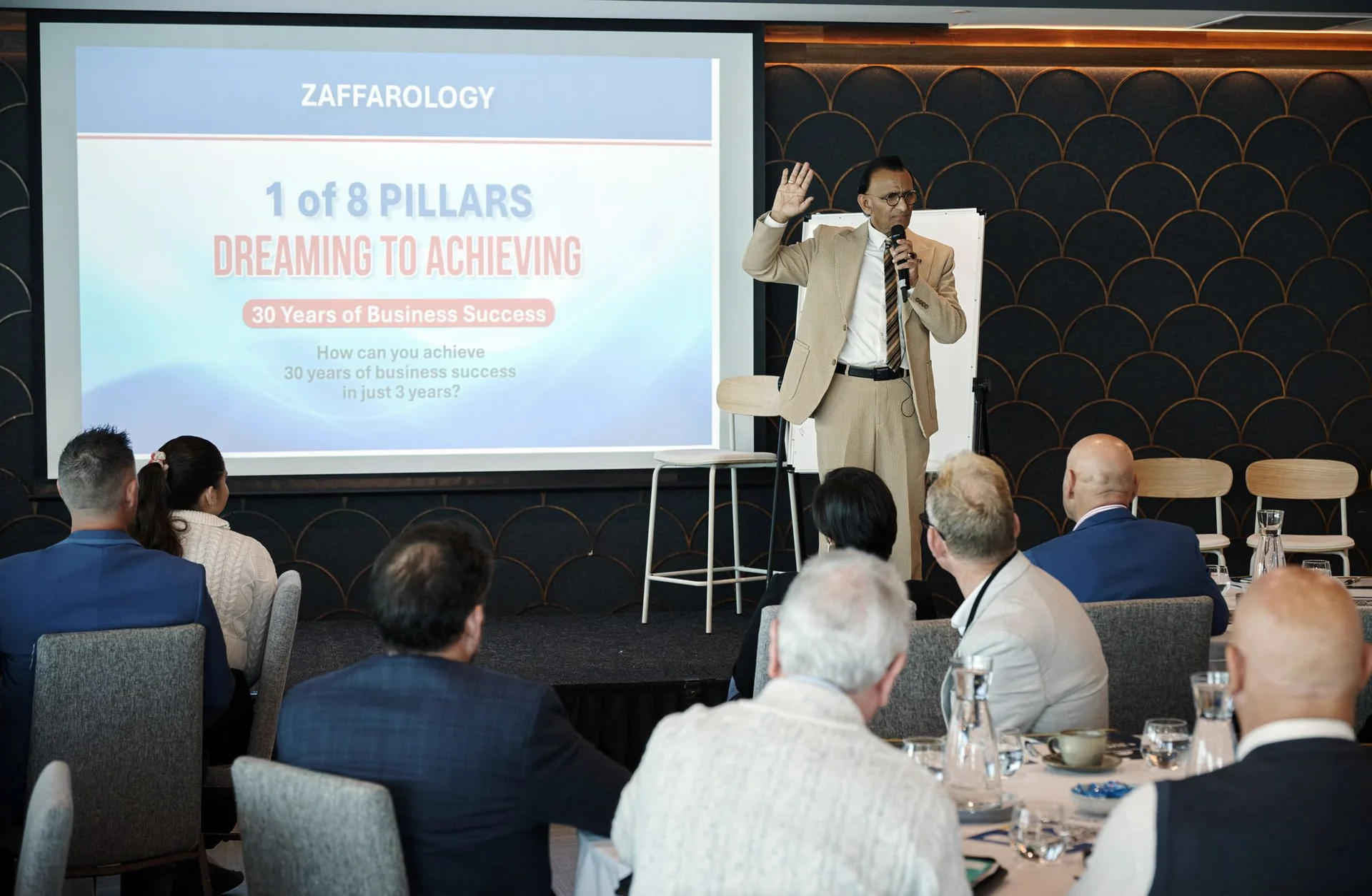 Zaffar Khan with a hand raised mid sentence in front of the Success Pillars slide