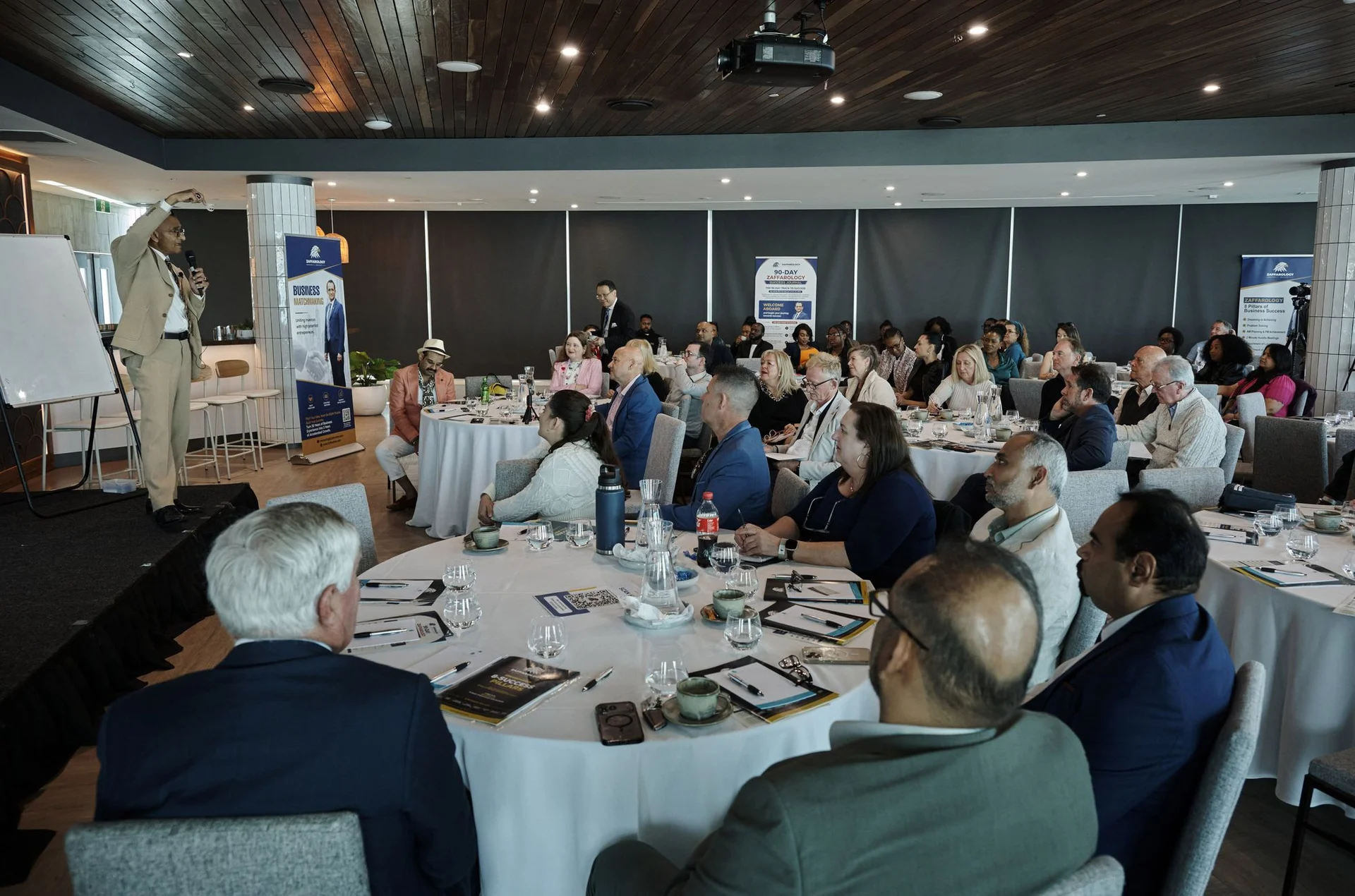 Zaffar Khan addressing the room from the stage beside a Business Matchmaking banner