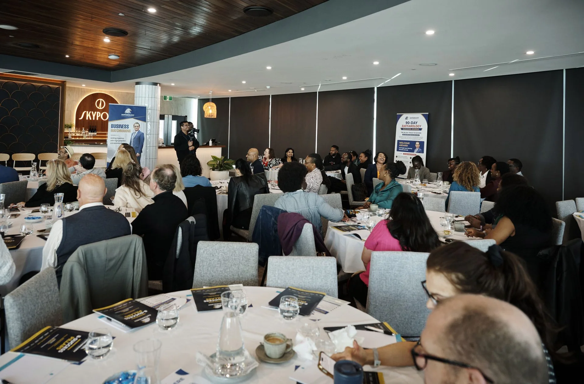 The room mid session with attendees at round tables and the SkyPoint bar behind